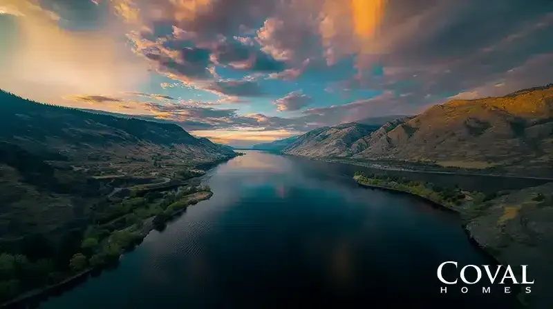 An elevated view of a wide, calm river flowing through a dramatic, steep mountain gorge. The rugged hills on the right are bathed in warm golden sunlight, while the left bank remains in deep shadow. Above, a spectacular sunset sky displays vibrant hues of orange, pink, and teal, which are perfectly reflected in the smooth, dark water below. The logo "COVAL HOMES" is displayed in the bottom right corner.