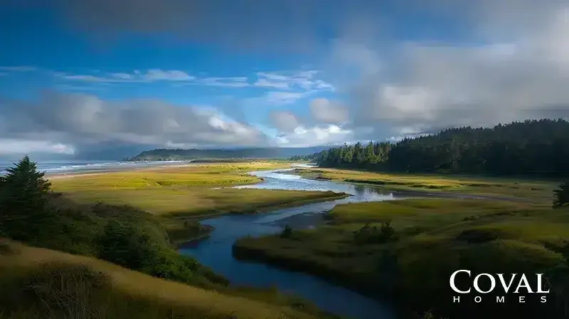 A scenic coastal landscape featuring a winding river meandering through a vibrant green and golden marshland toward a sandy ocean beach in the distance. To the right, a dense, dark evergreen forest borders the estuary. The scene is illuminated by patchy sunlight breaking through a dramatic sky filled with deep blue patches and low, heavy grey and white clouds. The logo "COVAL HOMES" is displayed in the bottom right corner.