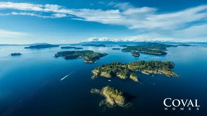 An aerial view of a picturesque archipelago featuring numerous lush, green, forested islands scattered across a vast expanse of deep blue water. A small white boat leaves a visible wake as it navigates the channels between the islands. In the distance, a hazy mountain range sits on the horizon beneath a bright blue sky filled with sweeping, wispy white clouds. The logo "COVAL HOMES" is displayed in the bottom right corner.