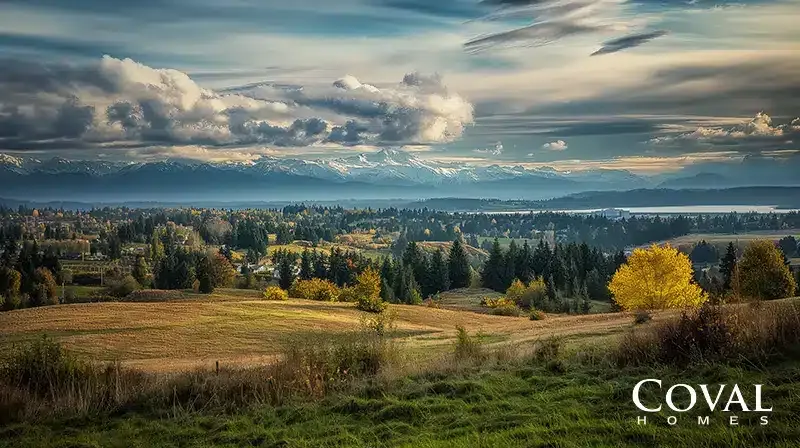 An expansive landscape view from a grassy field, looking over a sprawling valley filled with dark evergreen forests, patches of bright yellow autumn trees, and scattered suburban neighborhoods. Beyond the valley, a large body of water stretches horizontally across the middle distance, backed by a stunning, rugged range of snow-capped mountains spanning the horizon. The scene is set under a dramatic, heavily clouded sky with contrasting light and shadows cast over the terrain. The logo "COVAL HOMES" is displayed in the bottom right corner.