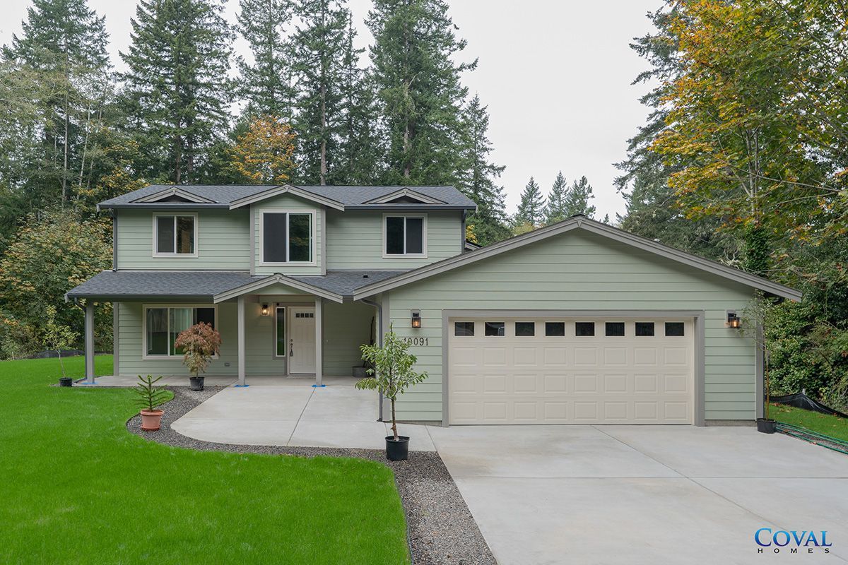 Two-story gray house with attached garage, front porch, and large driveway surrounded by trees