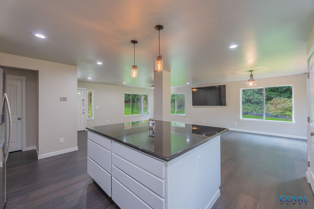 Modern kitchen with large island, dark countertops, pendant lights, and open living area with windows and TV