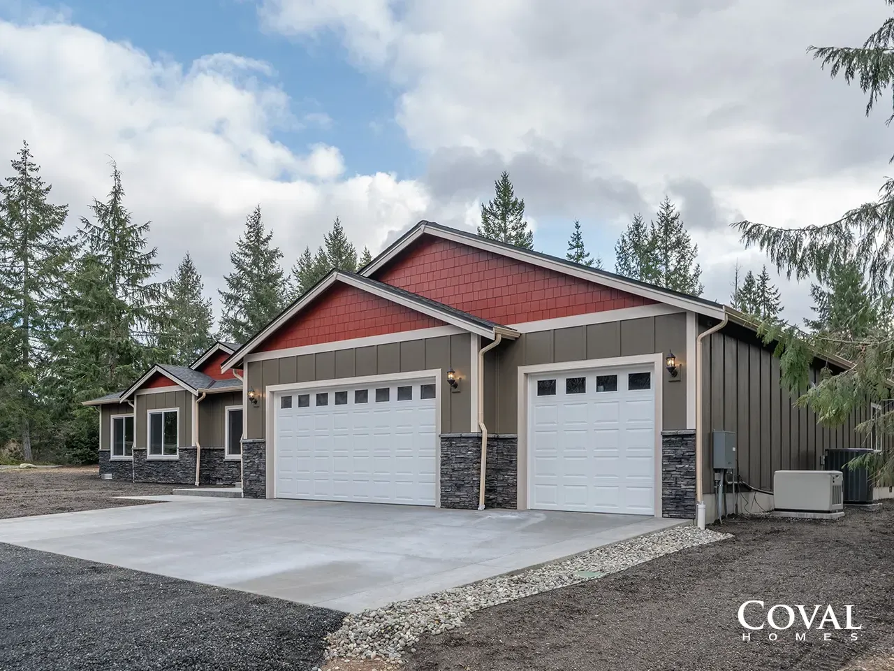 Modern three-car garage with beige siding, red roof accents, and stone trim on a gravel lot under cloudy skies