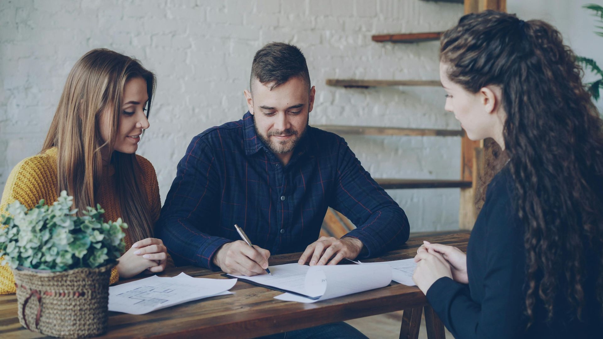 Three people discussing documents at a table in a bright office, with a laptop and plant nearby.