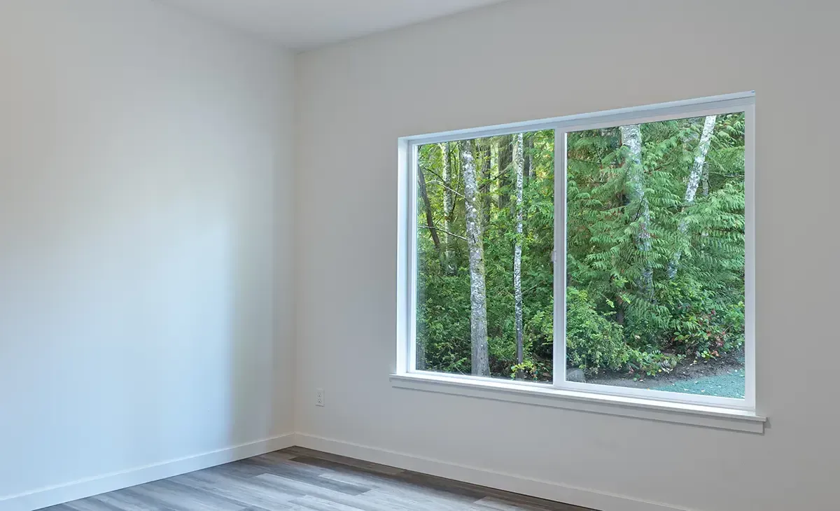 Empty white room with a large window overlooking green trees and sunlight.