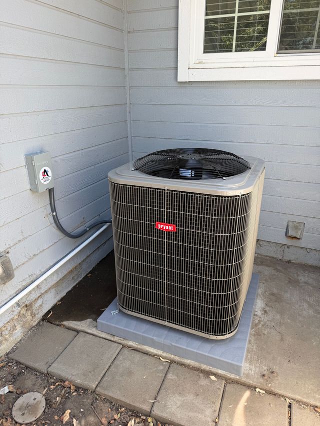 A light gray outdoor residential air conditioning unit sits on a concrete pad against a beige exterior wall.