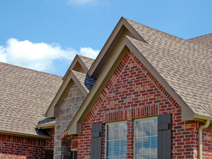 Brick house roof with gabled peaks, tan shingles, and blue sky with clouds above