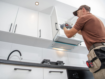 Person installing a kitchen range hood under white cabinets, using a drill in a modern kitchen