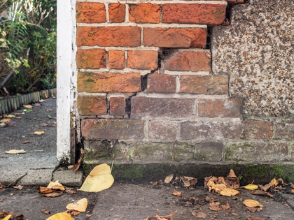 Damaged brick wall corner with cracked mortar and damp lower bricks by a wet sidewalk