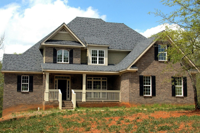 Two-story brick house with a front porch, gray roof, and red flowers in the yard