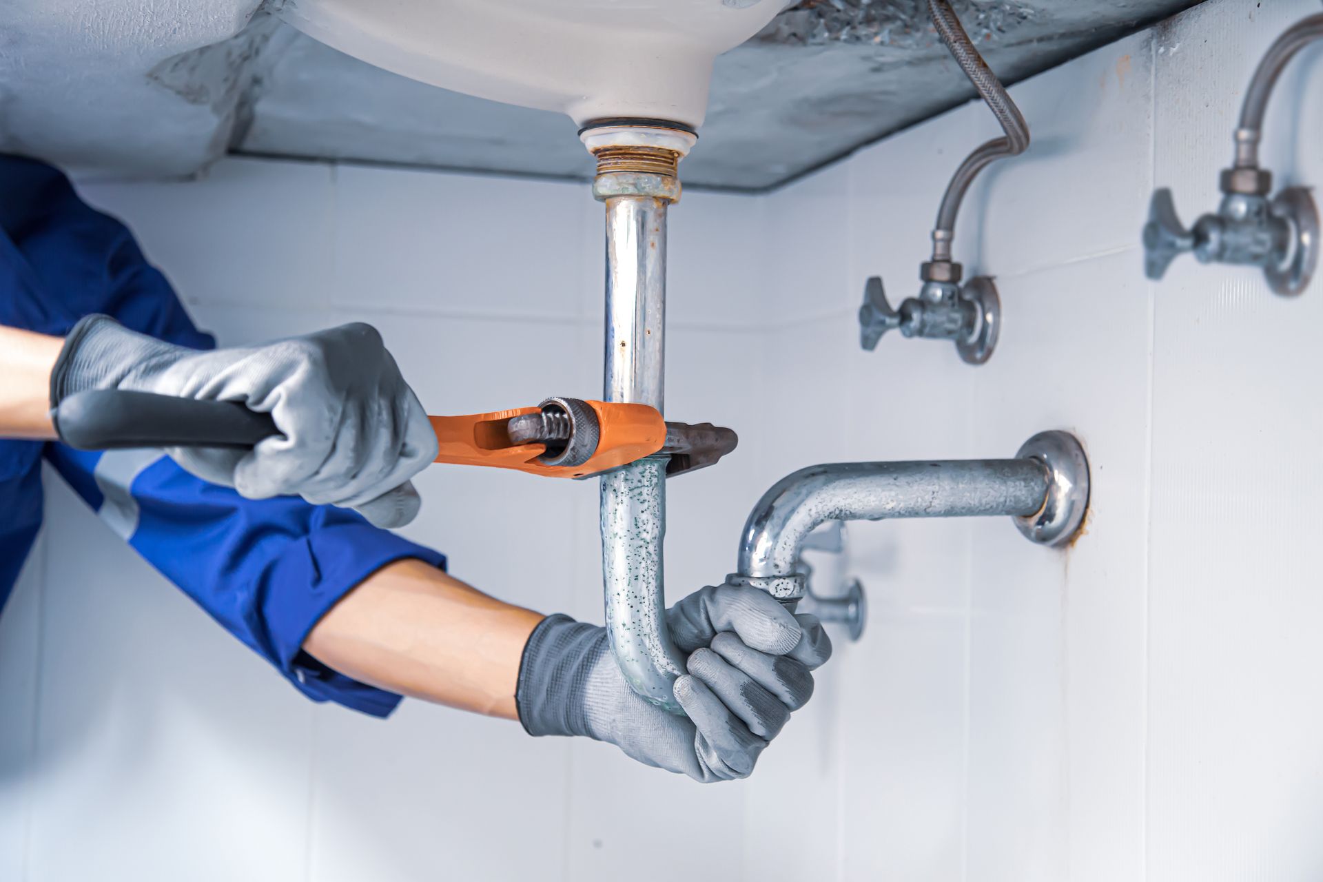 A person in a blue uniform and gloves uses a wrench to repair a silver metal drain pipe under a sink. A person in a blue uniform and gloves uses a wrench to repair a silver metal drain pipe under a sink.