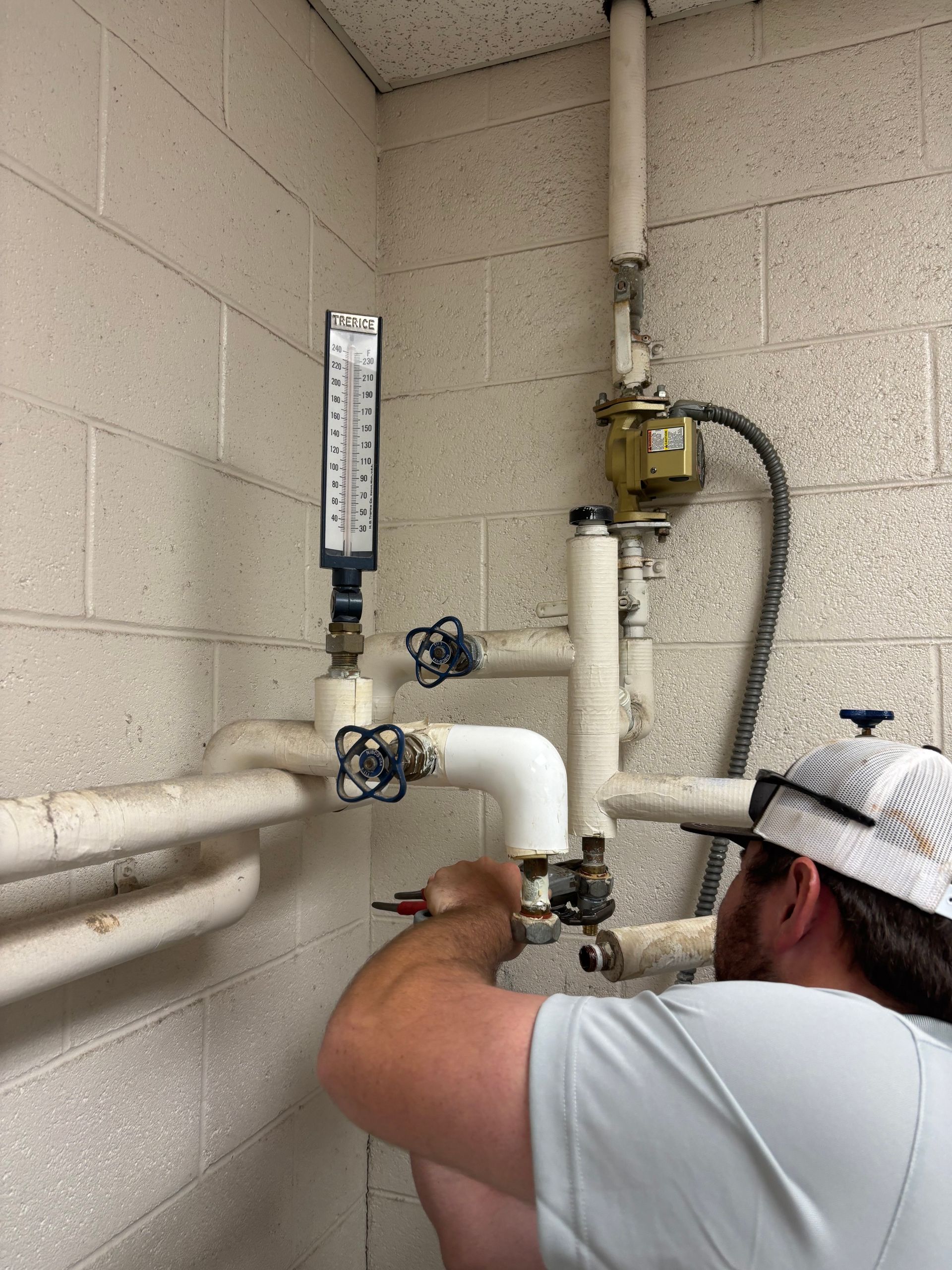 Worker inspecting basement plumbing with white pipes and valves on cinderblock walls.