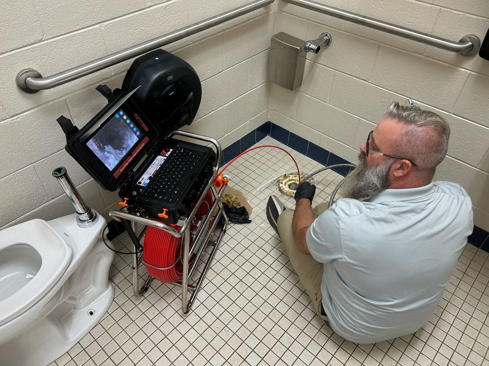 Technician inspecting a pipe with a camera in a small restroom, seated on the floor beside a monitor