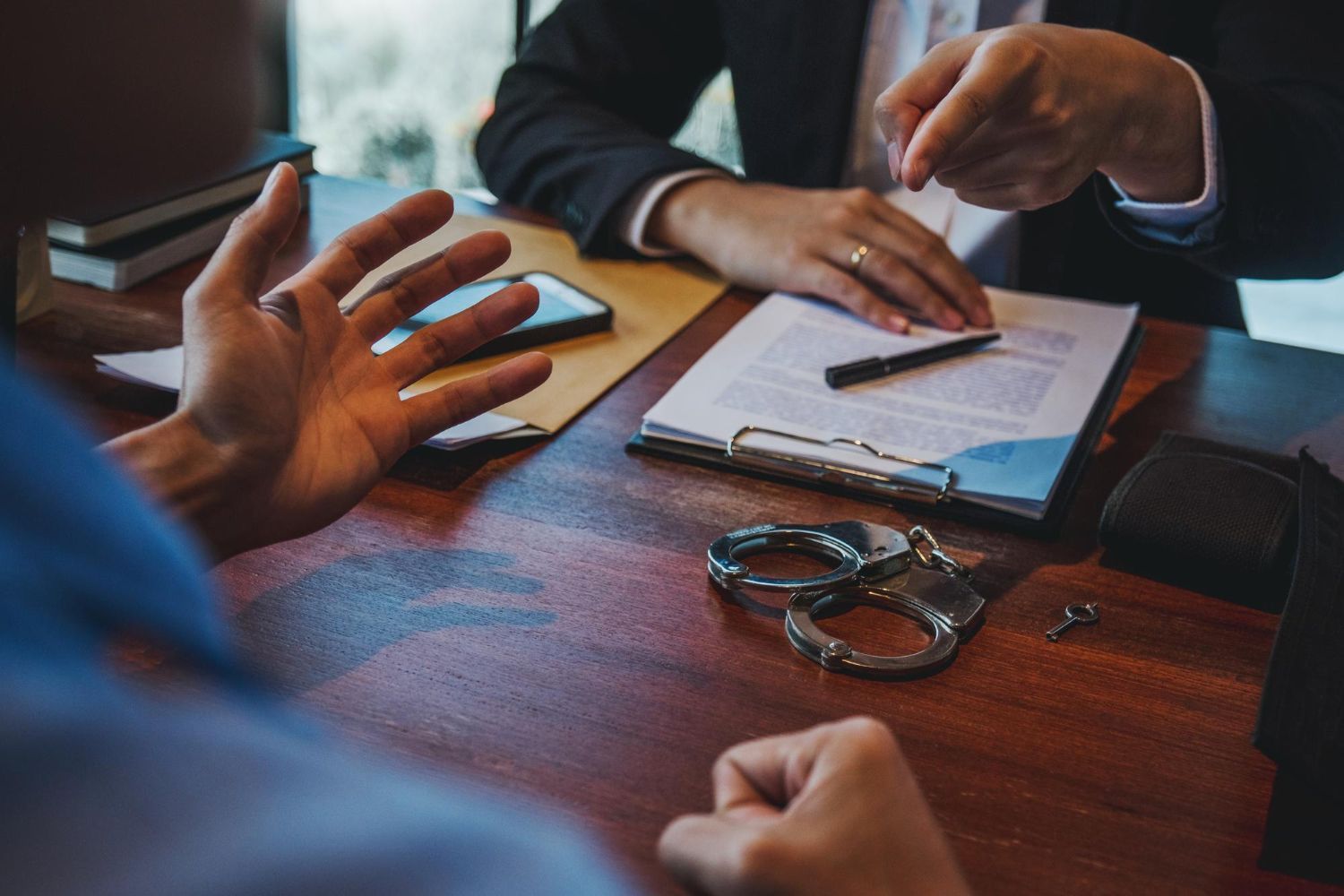 Two people in a meeting discussing paperwork at a wooden desk with a notebook and handcuffs.