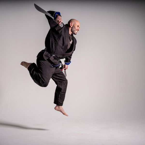 Two martial artists mid-air in a studio, one delivering a flying kick against a gray background.