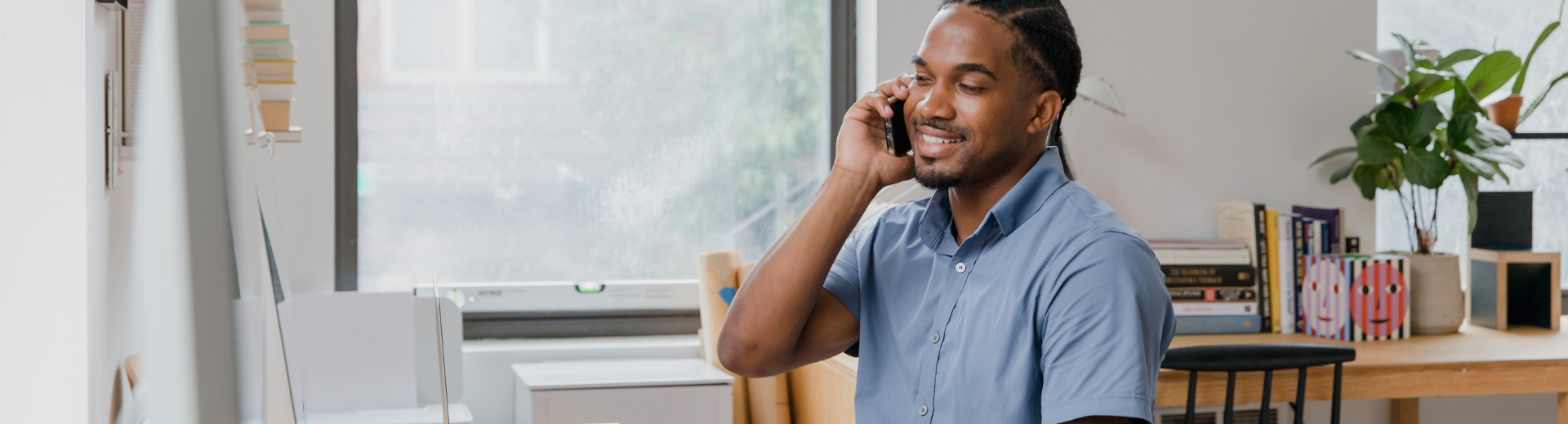 Homem sorrindo enquanto fala ao telefone dentro de casa, perto de uma janela, com luz natural.