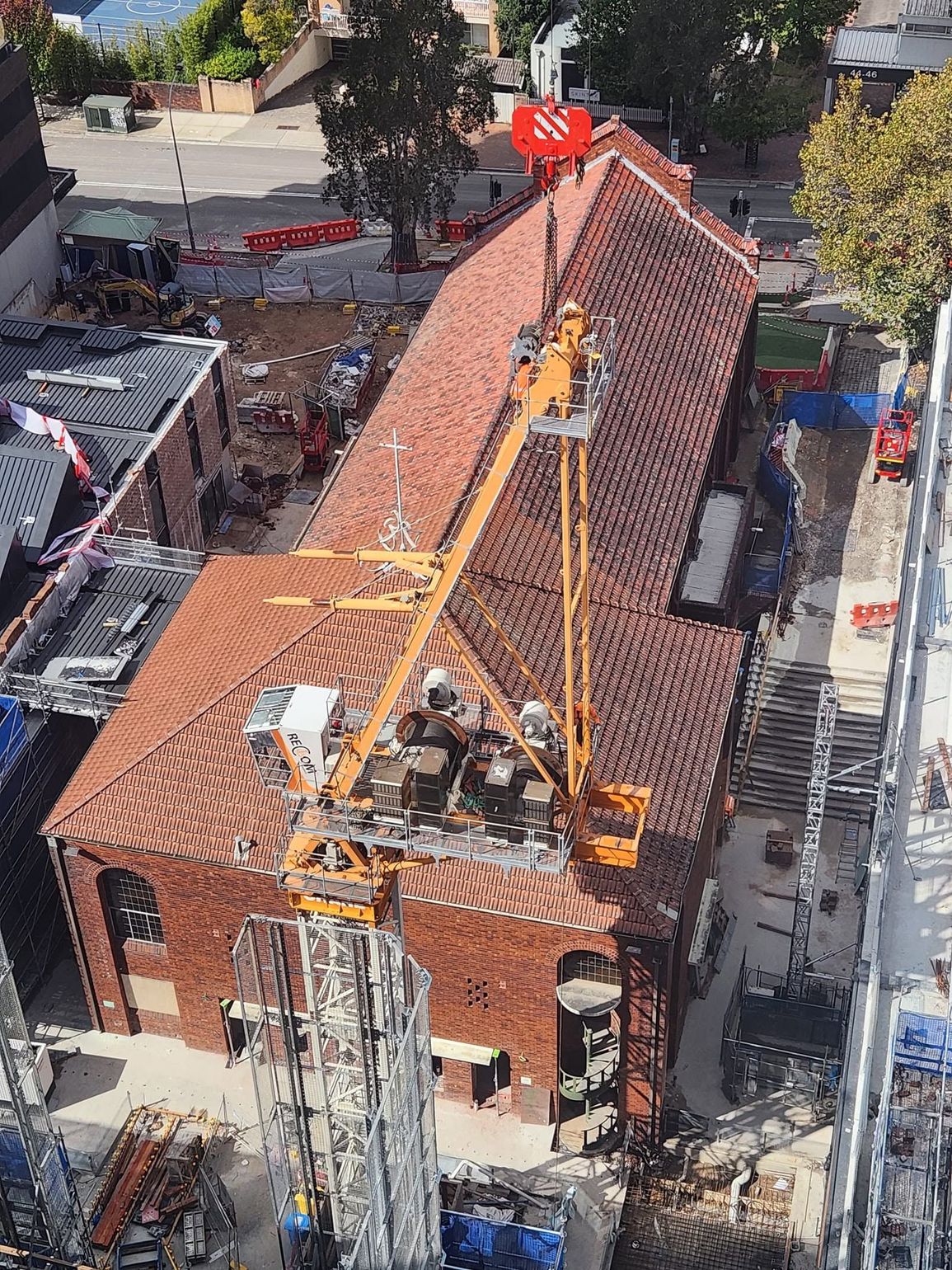 Brick building under construction with a red-tiled roof, crane, and workers on top.