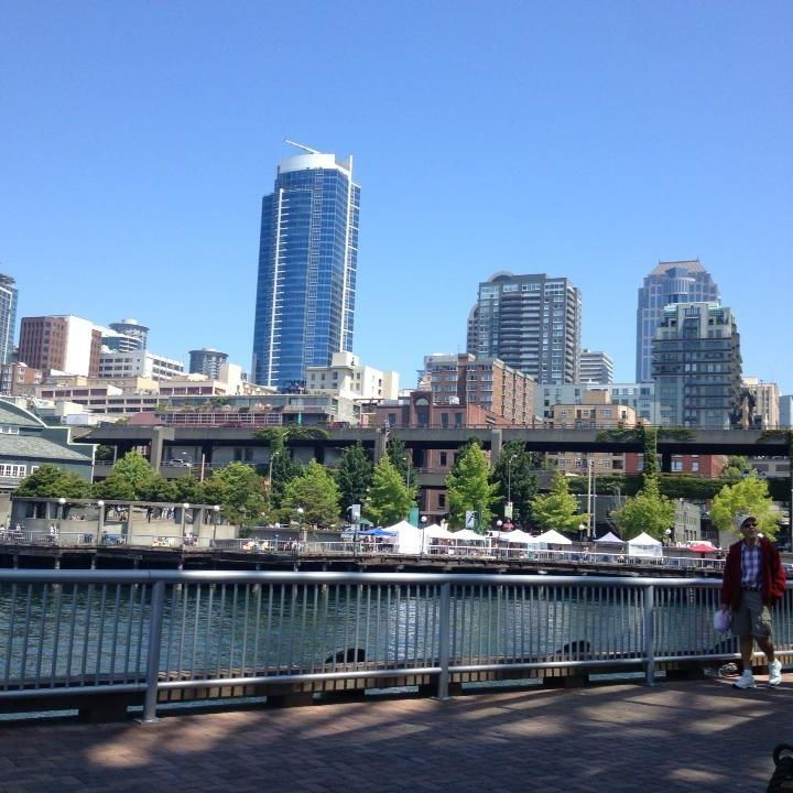 Seattle waterfront with city skyline; man in jacket stands near railing, sunny day.