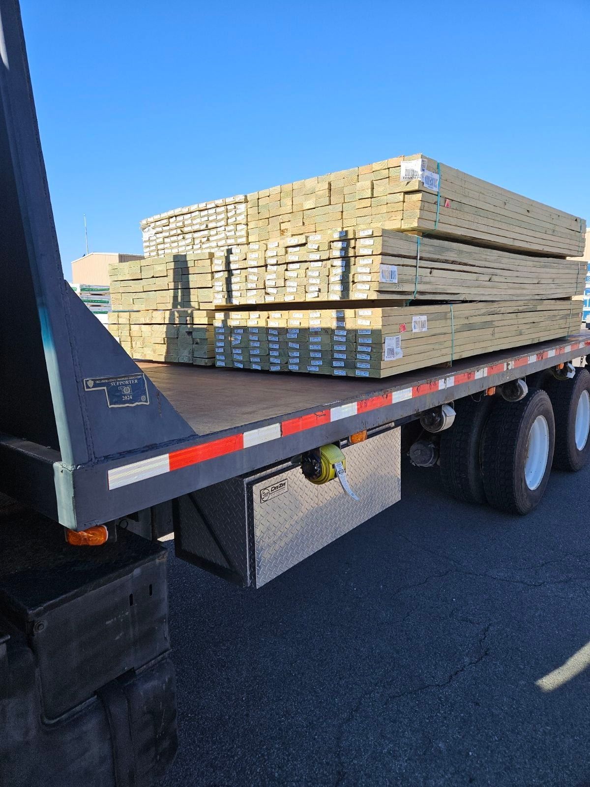 Wooden lumber stacked on a flatbed truck, outdoors. The lumber is light brown and secured.