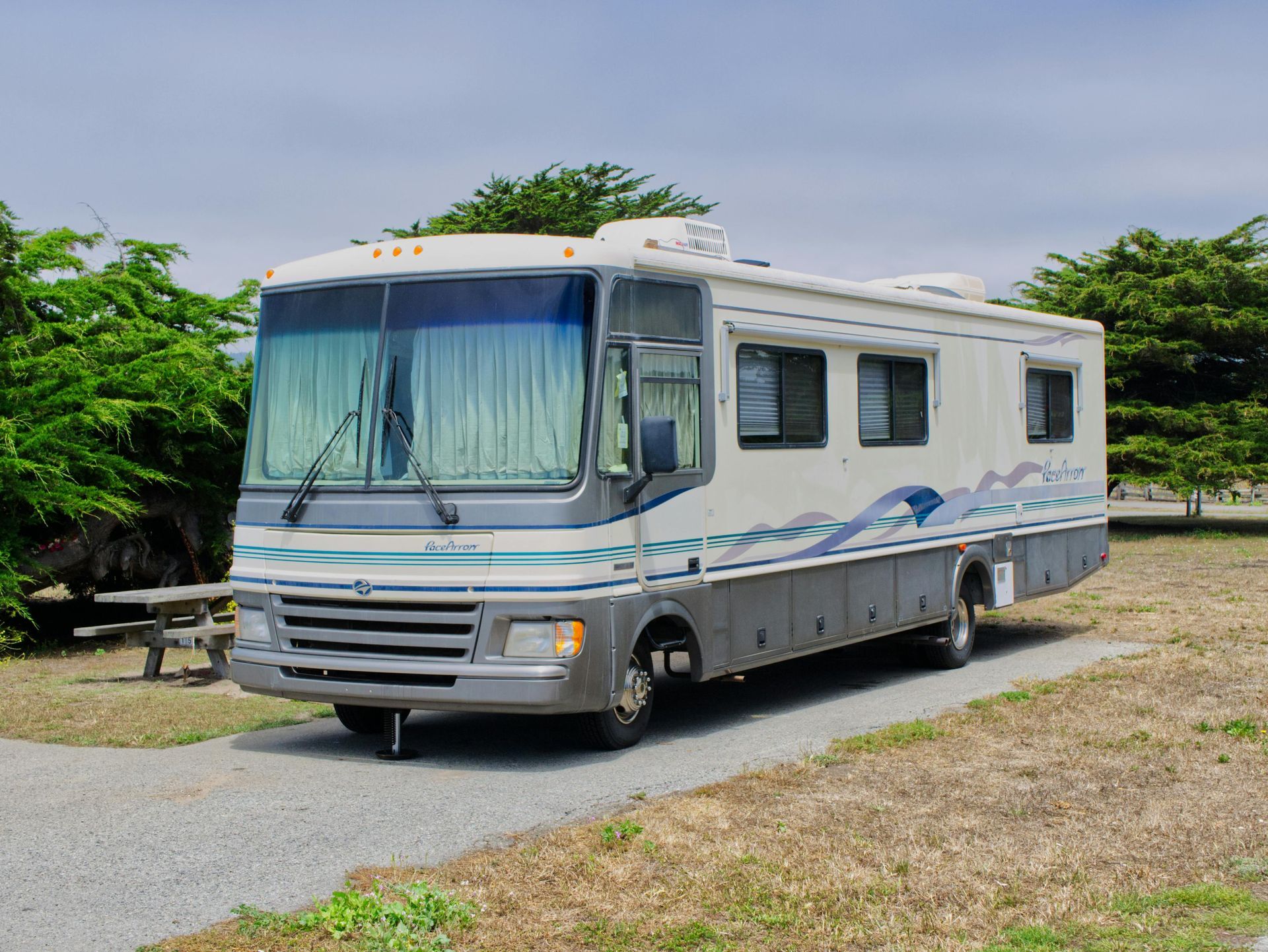 RV parked on a gravel pad with a picnic table, against a backdrop of green trees and a cloudy sky.