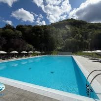 Piscina all'aperto con acqua turchese, lettini, ombrelloni e verdi colline sotto un cielo nuvoloso.