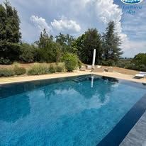 Piscina in giardino con acqua azzurra, terrazza beige, alberi e cielo nuvoloso.