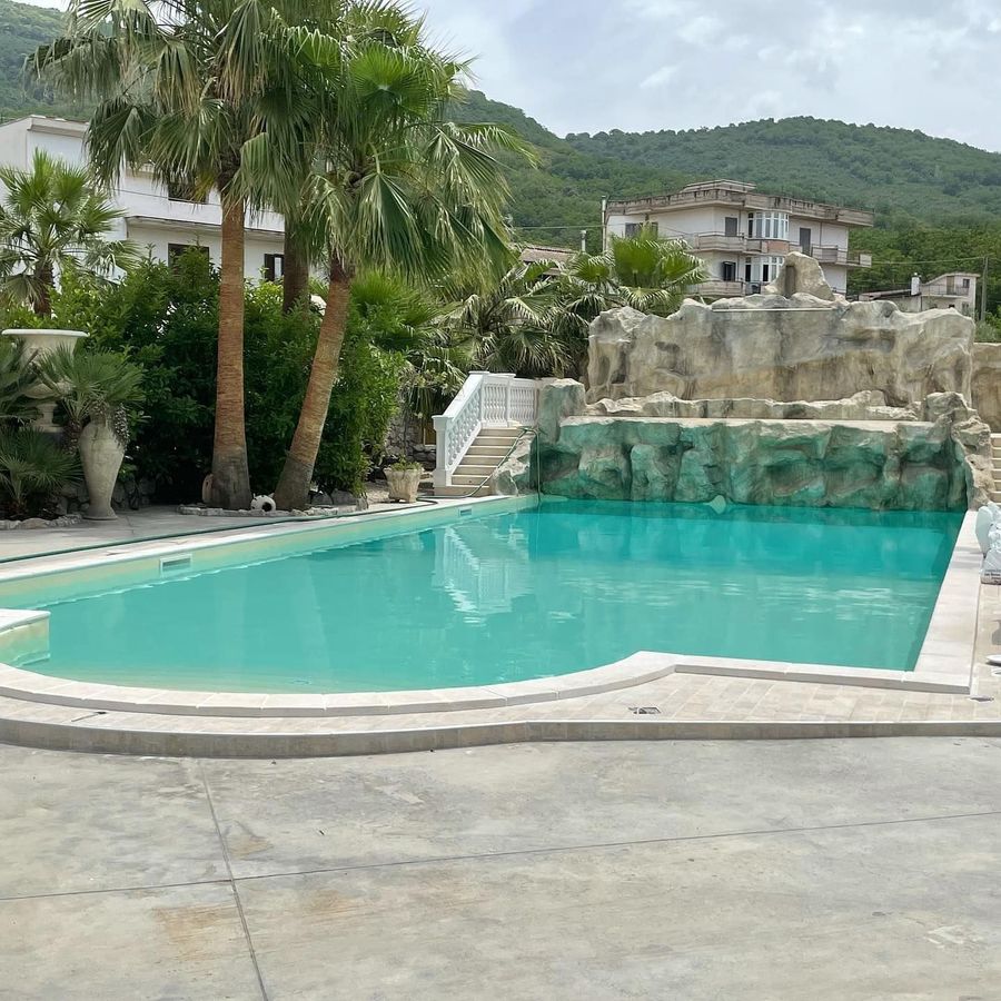 Piscina turchese accanto a palme e a una parete rocciosa, con case sulla collina sullo sfondo.