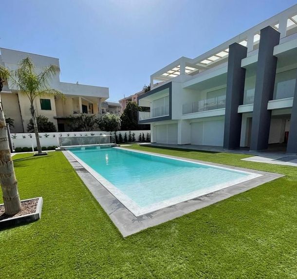 Cortile di una villa moderna con piscina rettangolare turchese, prato verde ed edifici bianco-grigi.