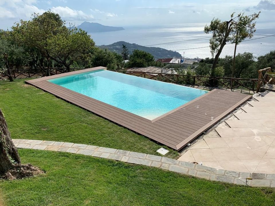 Piscina all'aperto con acqua turchese su una terrazza erbosa con vista panoramica sulle montagne e sul mare.