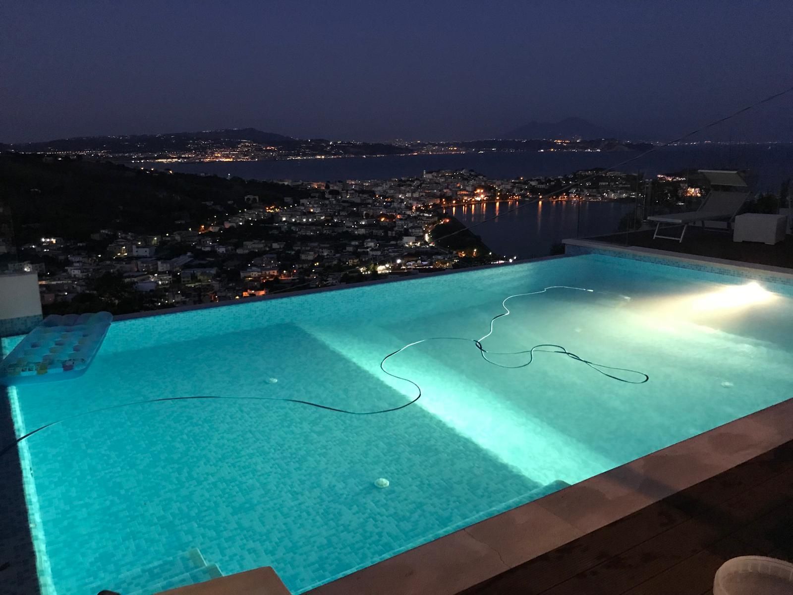Piscina a sfioro illuminata al crepuscolo con vista sullo skyline della città e sulle colline in lontananza.