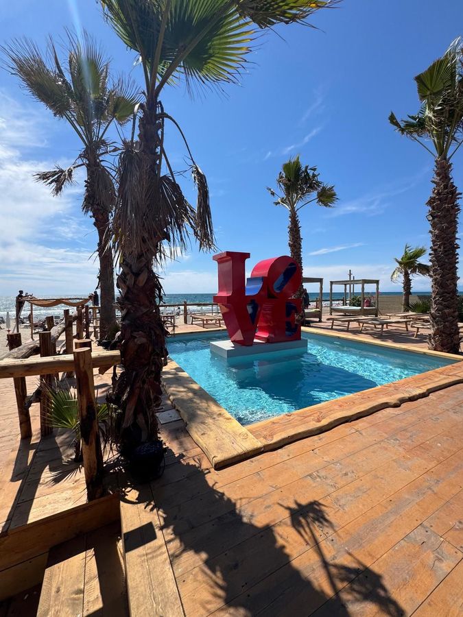 Patio a bordo piscina con palme e una grande scultura rossa affacciata sull'oceano.