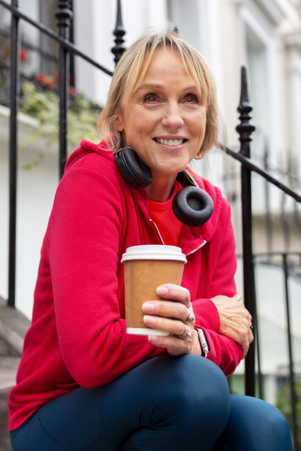 Smiling woman in a red sweater holding a coffee cup, seated outdoors with headphones around her neck