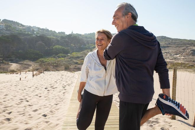 Two people smiling on a sunny beach, standing near dunes and holding a surfboard