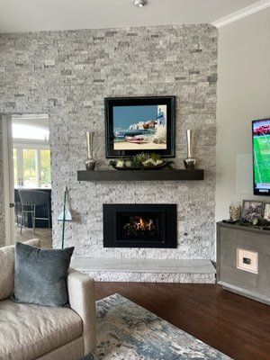 A fireplace with a black insert and floating mantel on a light stone wall, framed by a living room with a gray armchair.