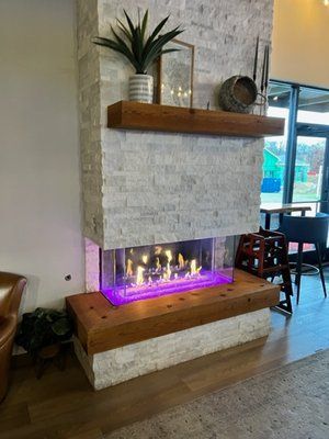 A modern stone fireplace with a wooden mantle and base, featuring a see-through glass firebox with purple LED lighting.