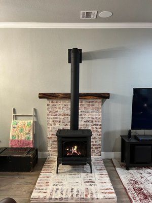 A lit black wood-burning stove sits on a white brick hearth in front of a matching brick wall with a rustic wood mantel.