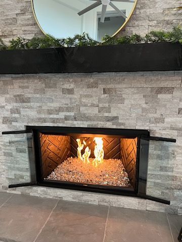 A lit fireplace with light-colored fire glass in a stone-tiled hearth, topped by a black mantle and circular mirror.