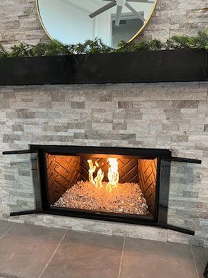 A lit fireplace with glass rocks inside a light-colored, stacked stone wall, topped with a black mantel and round mirror.
