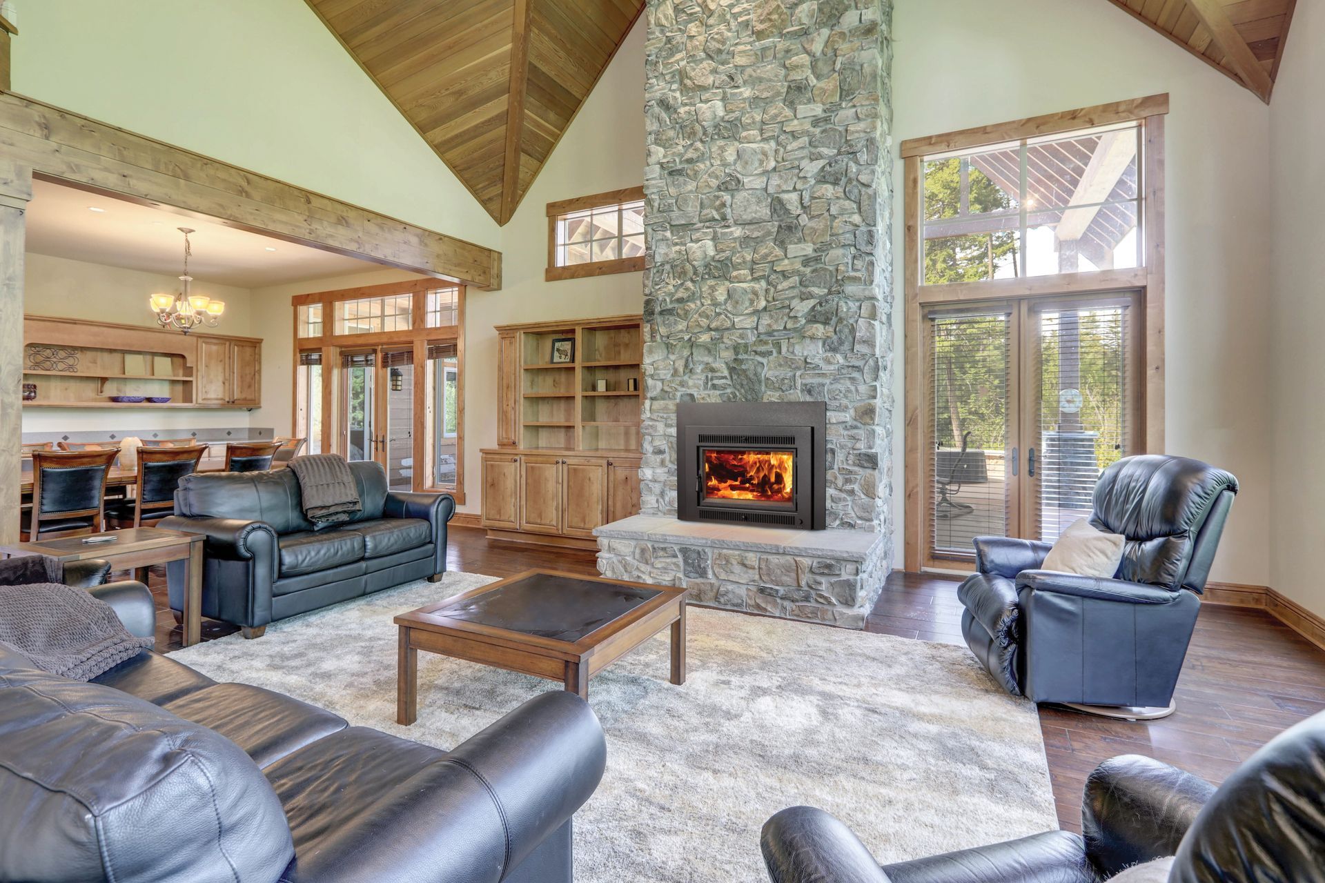Living room with a stone fireplace, leather sofas, a rug, and a wood-paneled ceiling, opening into a dining area.
