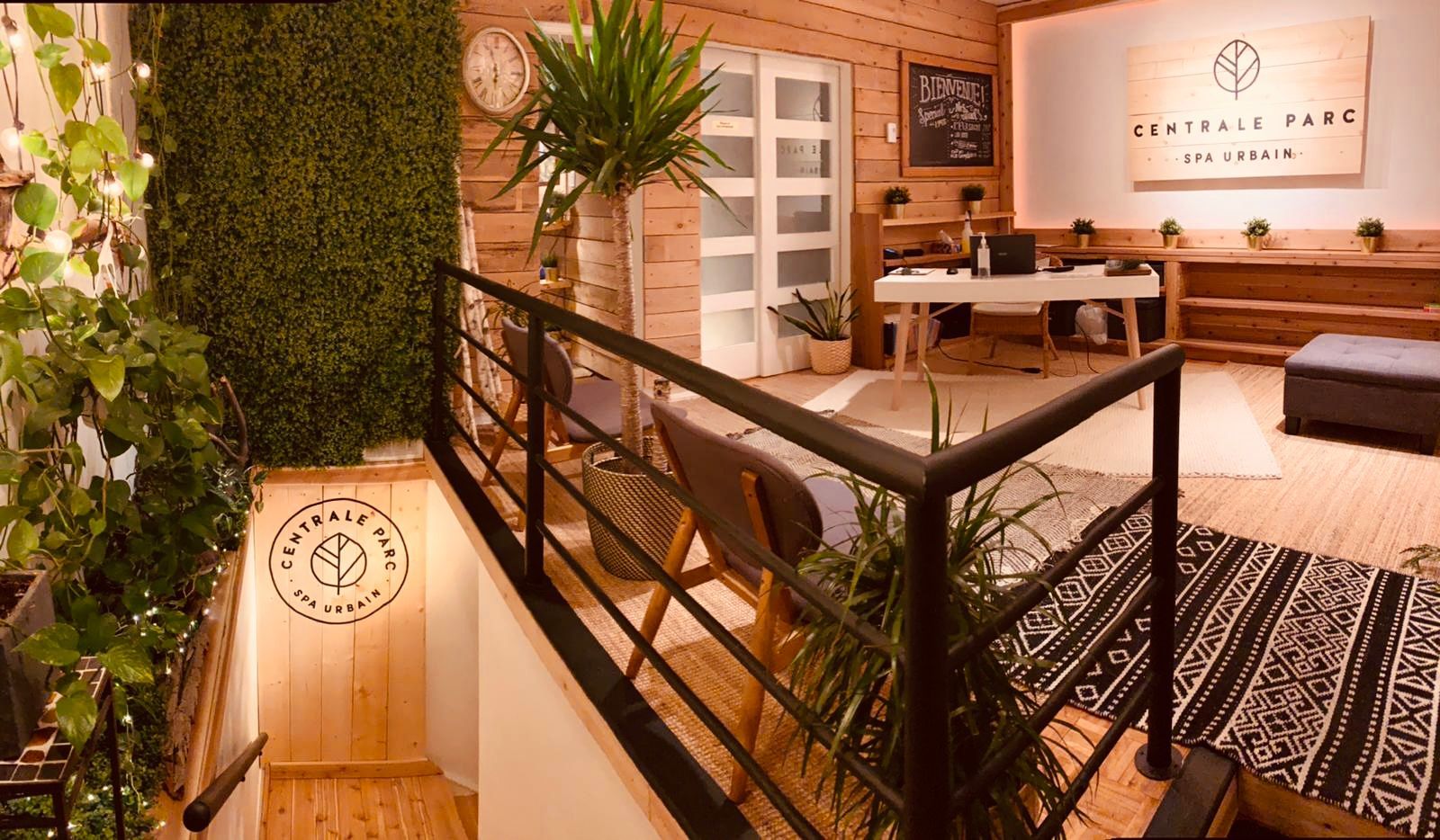La réception du Spa Centrale Parc comprend un bureau d'accueil, une grande enseigne murale, des étagères en bois et une décoration végétale dans une ambiance apaisante.
