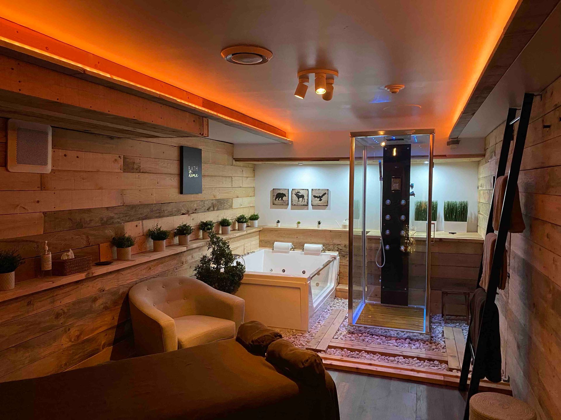 A dimly lit spa room features a massage table, a white armchair, a jetted tub, and a glass-enclosed shower against wood walls.