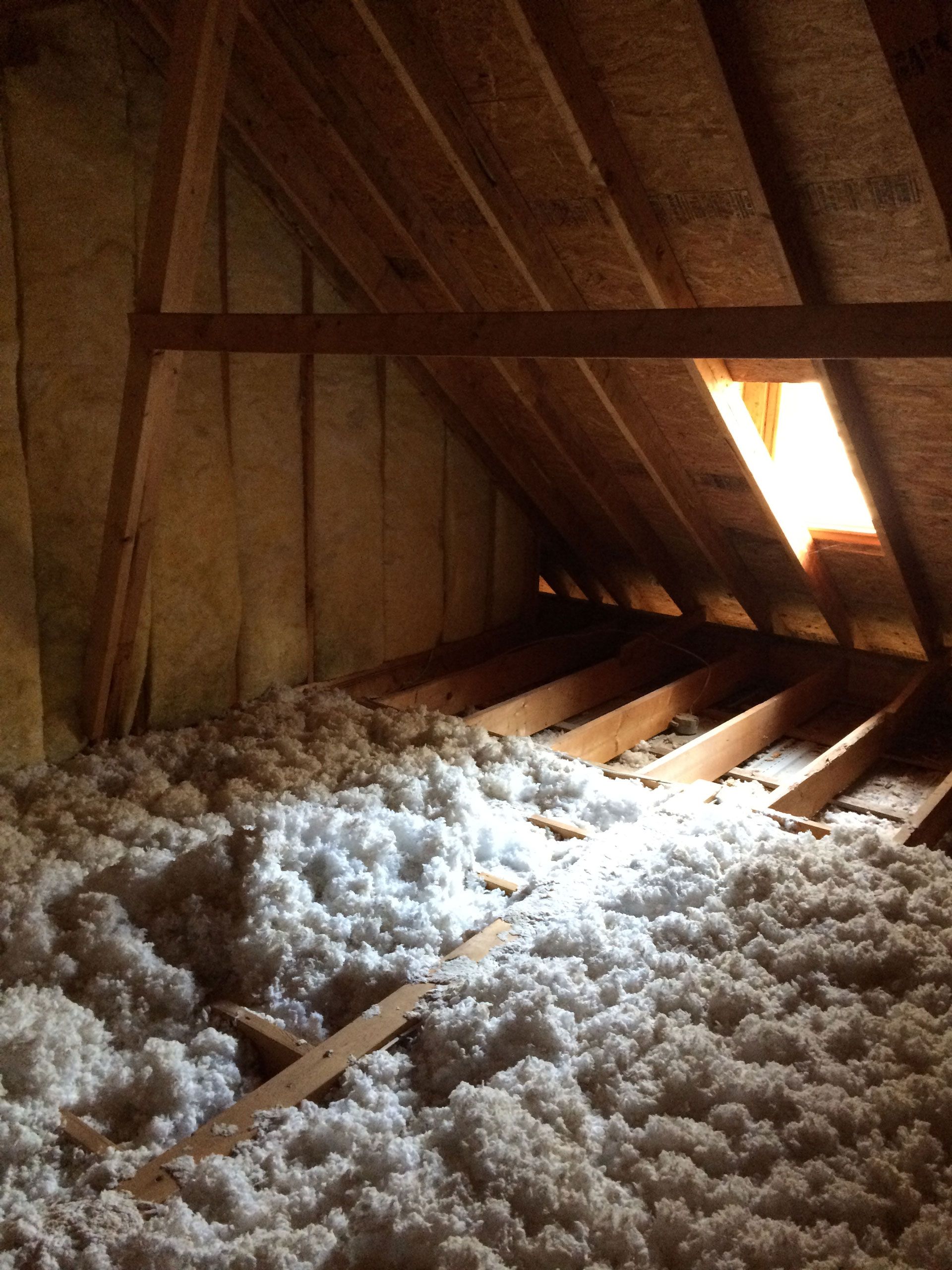 Attic interior with cellulose insulation covering the floor, wooden beams, and a small window emitting sunlight.