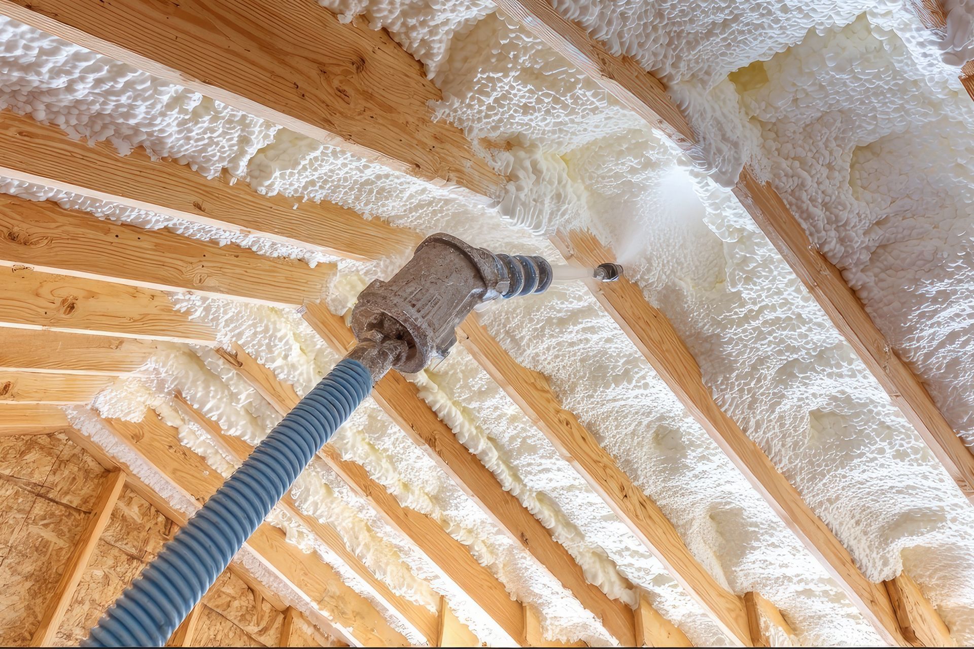 Spray nozzle applying white foam insulation between wooden roof rafters during construction.