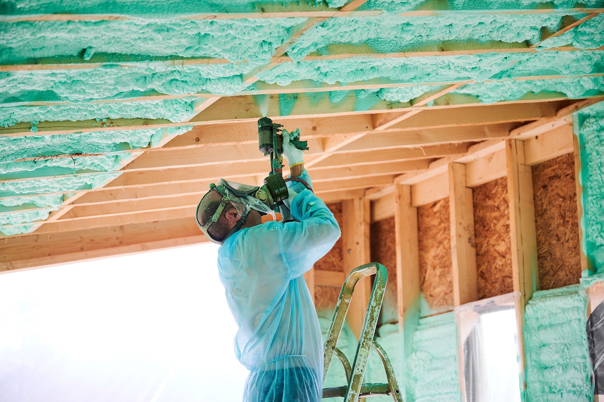 Worker installing insulation with a nail gun on a construction site. Green foam, wood framing, protective gear.