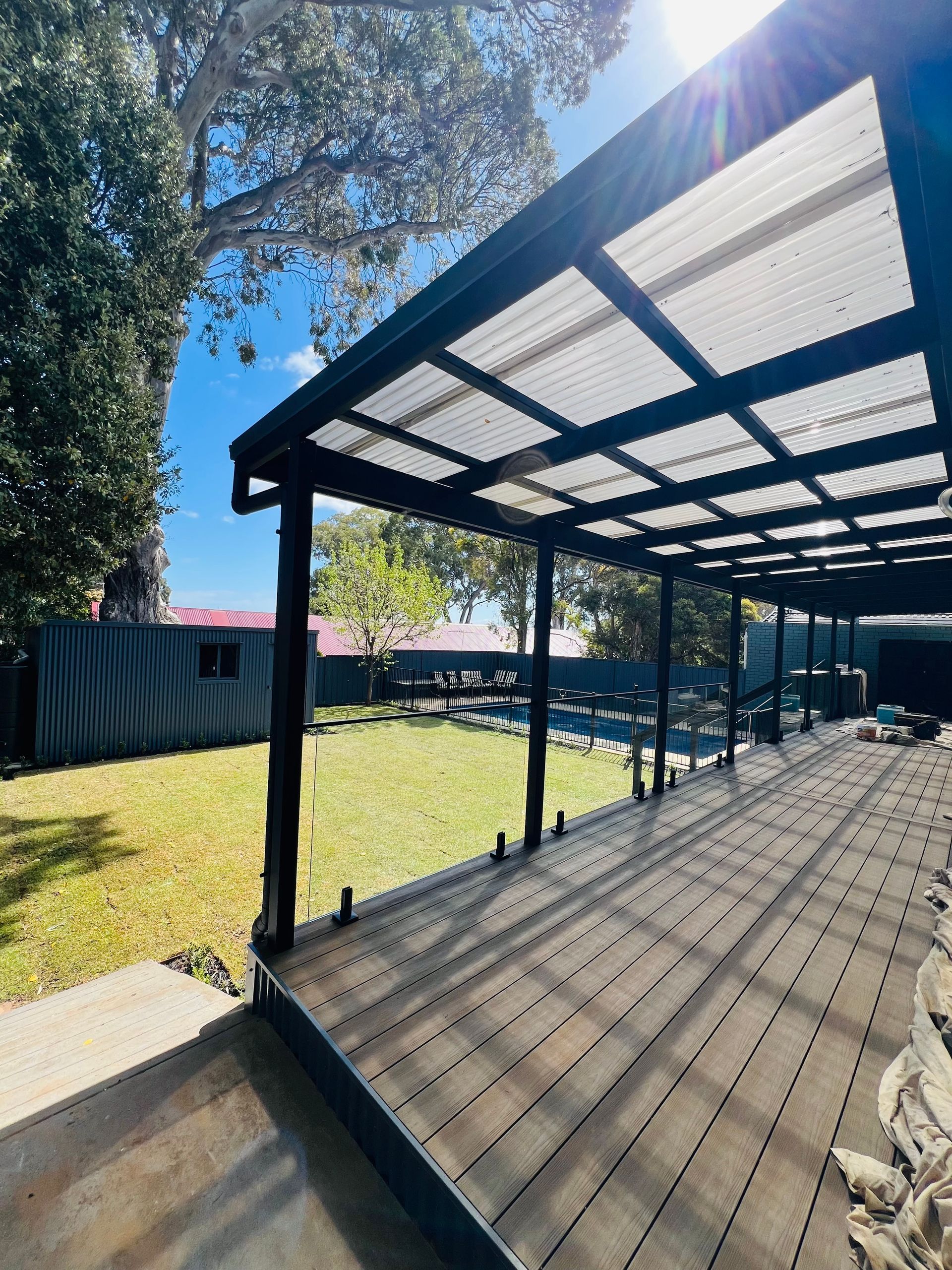 A brown wooden lattice pergola roof with a translucent corrugated covering and two ceiling fans against a brick wall.