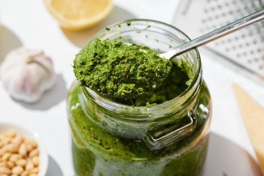 Barattolo di pesto verde con cucchiaio, circondato da aglio, limone e pinoli su una superficie bianca.