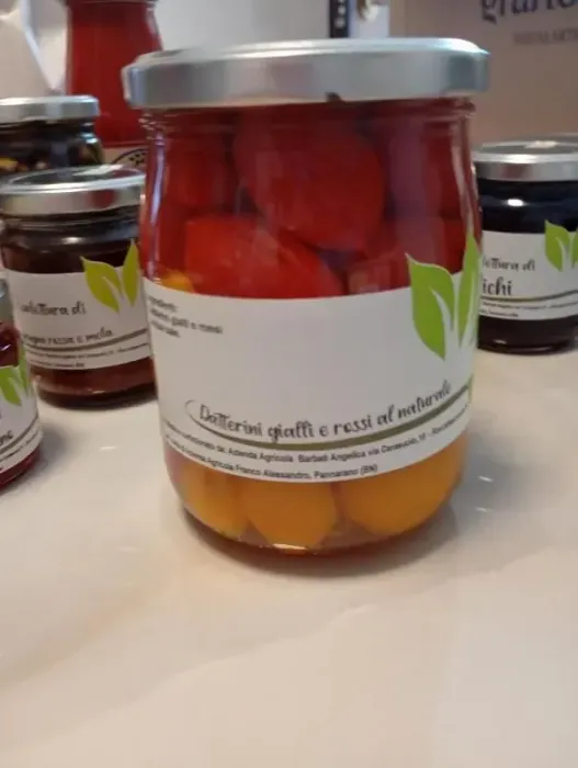 Su un tavolo si vedono dei barattoli di frutta sciroppata rossa e gialla, con altri barattoli sfocati sullo sfondo.