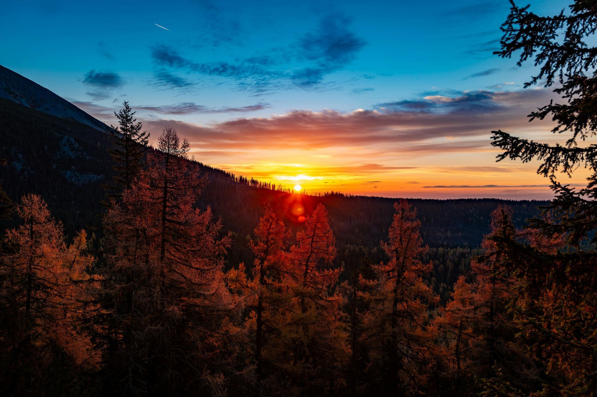 A vibrant sunset glows behind dark mountain silhouettes and autumnal orange forest trees under a deep blue sky.