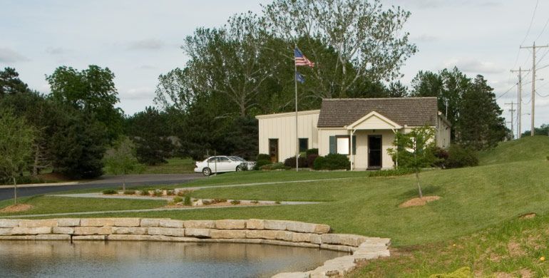 Small beige building by a pond with an American flag and grassy hill in a park setting