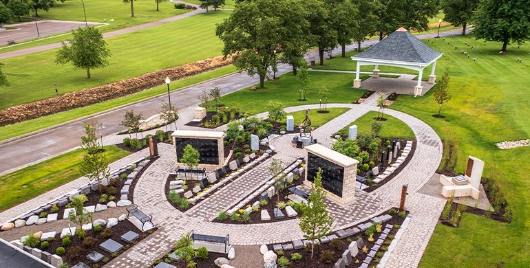 Aerial view of a stone memorial garden with curved paths, benches, and a white gazebo in a green park.